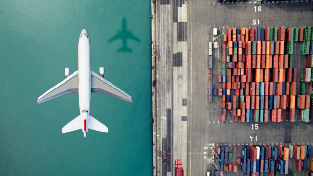 An overhead view of an airplane flying above cargo containers at a port