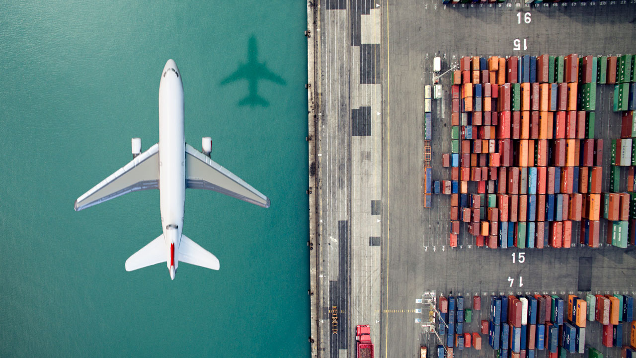 An overhead view of an airplane flying above cargo containers at a port