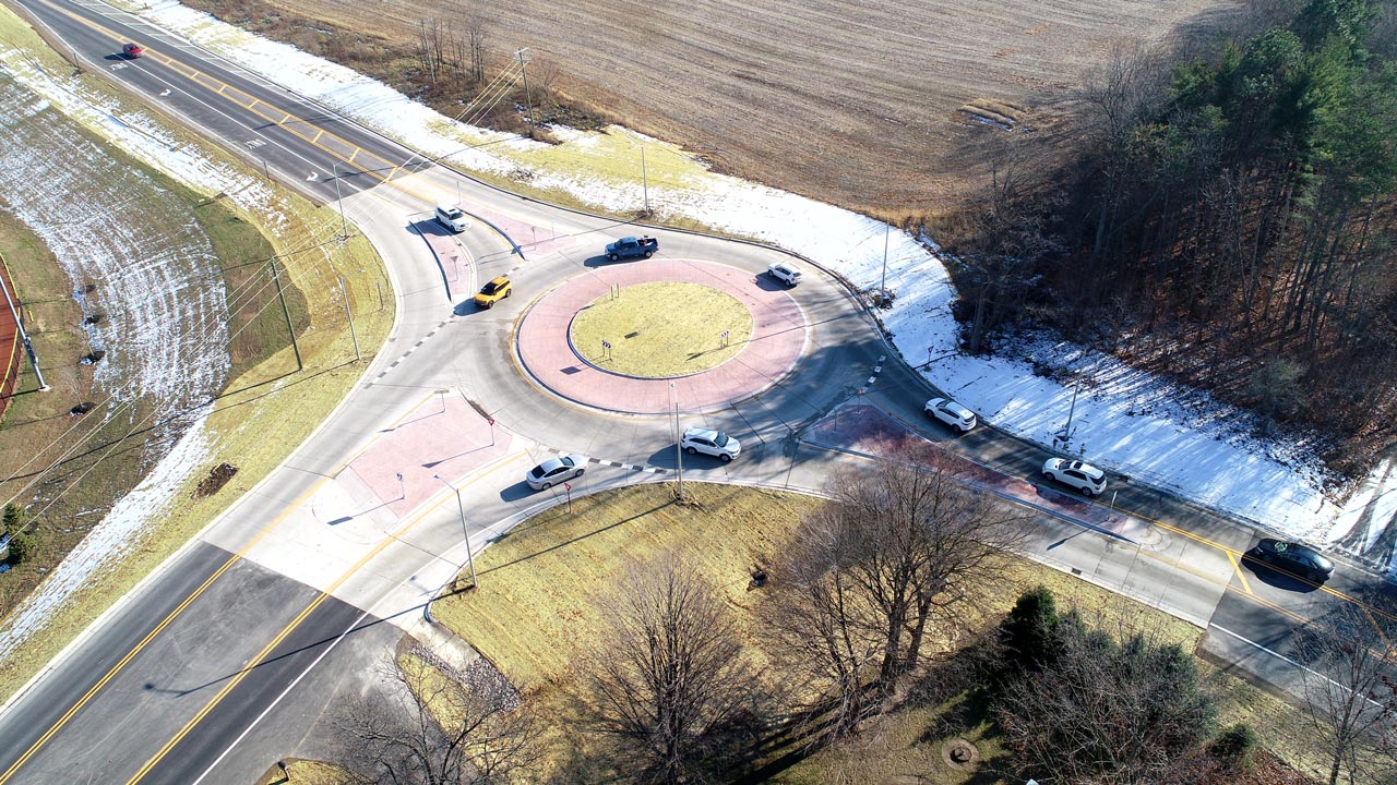 Aerial view of vehicles traveling around a roundabout surrounded by fields and partially melted snow