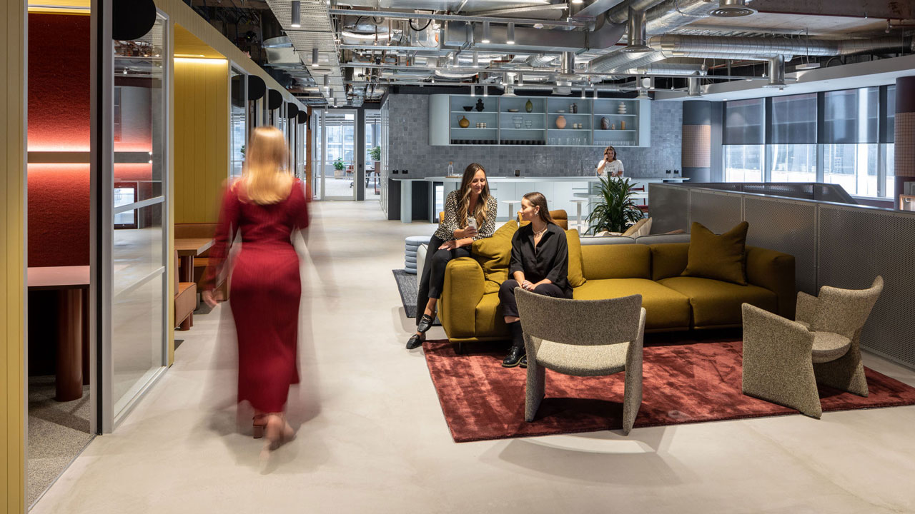 A woman walks through an open space with a mix of seating types, colorful accent walls and furniture, and exposed ductwork