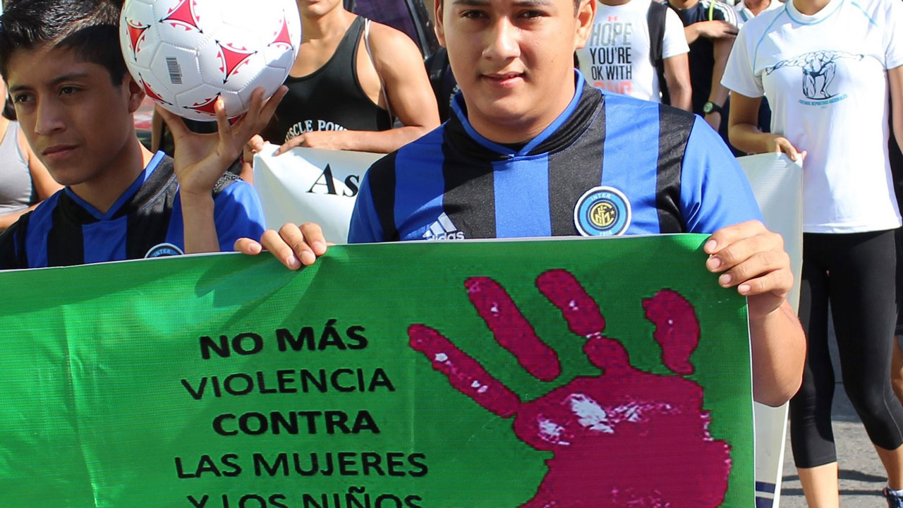 A boy walks and holds a green sign that reads “No more violence against women and children”