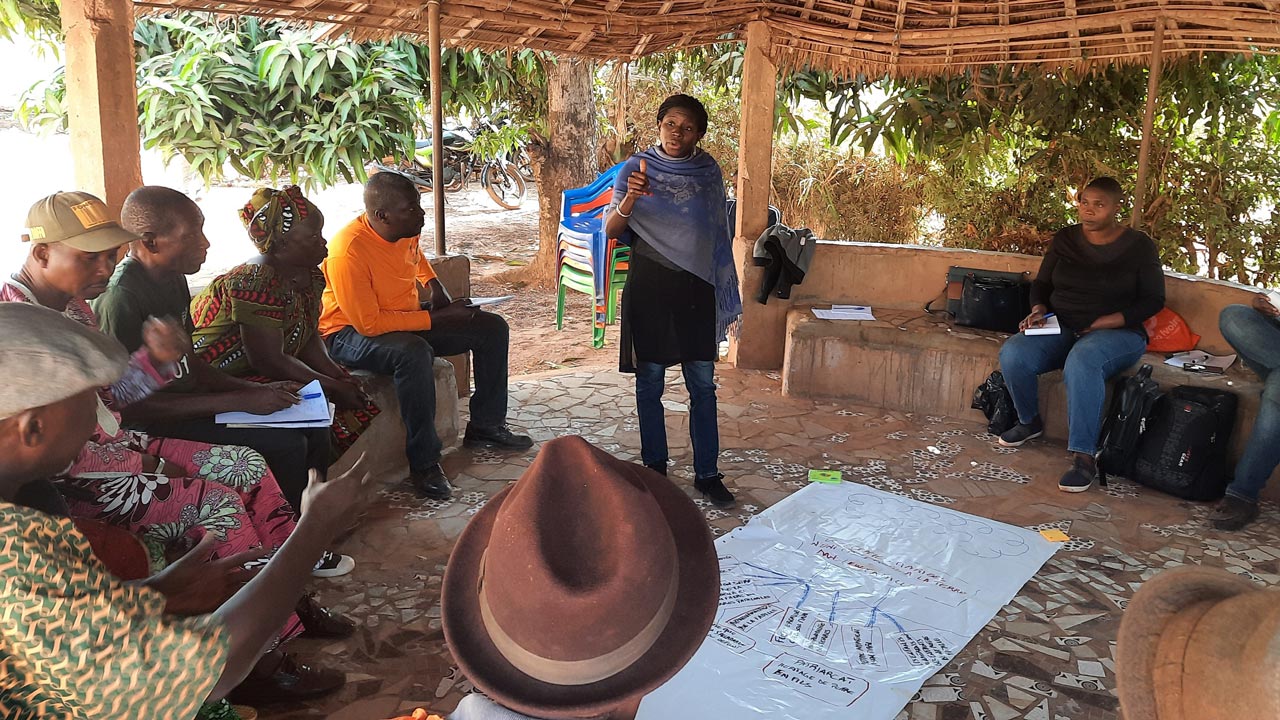 Woman in Cote d’Ivoire leads a community meeting standing over a large paper on the ground with a group of people in a circle looking at her