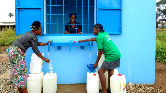 Two people filling jerrycans with water at a watering station with a smiling station attendant inside a booth