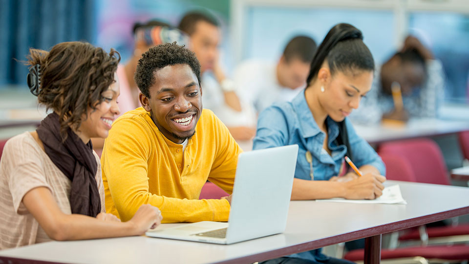 Three Black students sit at a table, with two of them working on a laptop, and the third writing on a piece of paper