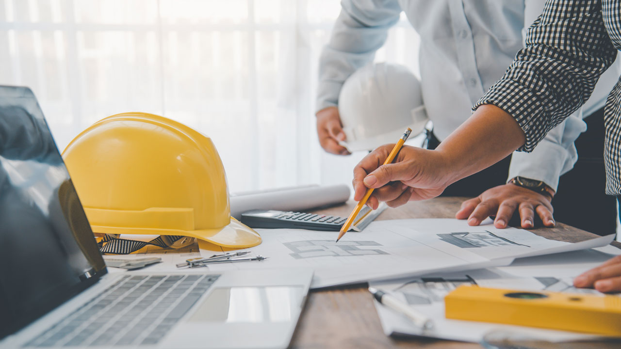 An open laptop, yellow hard hat, and design documents on a table with people gathered around reviewing the documents