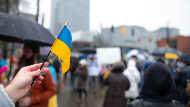 A hand flying a small flag of Ukraine against a blurred urban backdrop