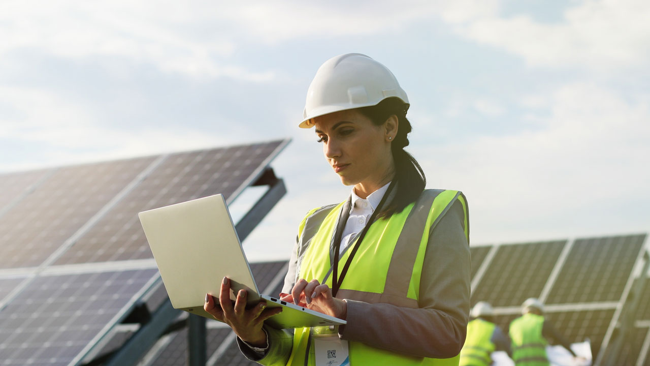 Female engineer in safety gear using laptop with solar panels in the background