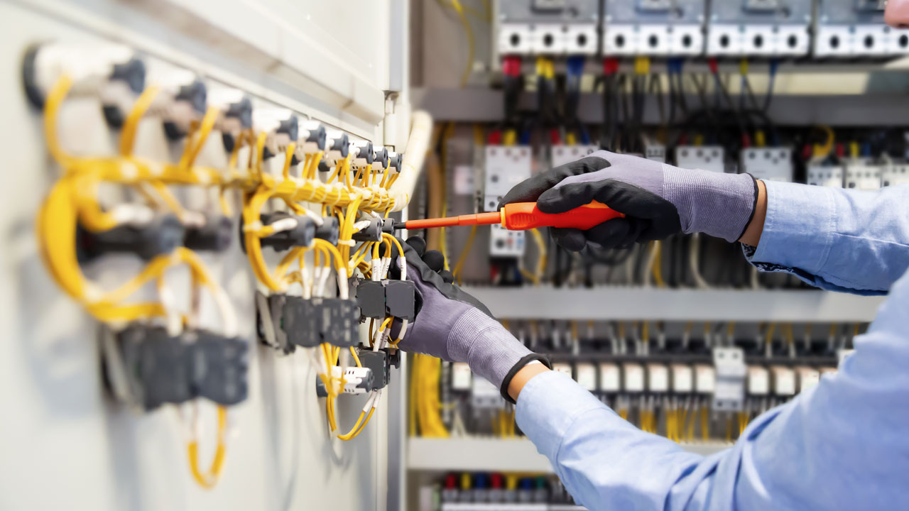 erson in gloves is using a screwdriver to connect yellow cables in a control panel filled with various electrical components
