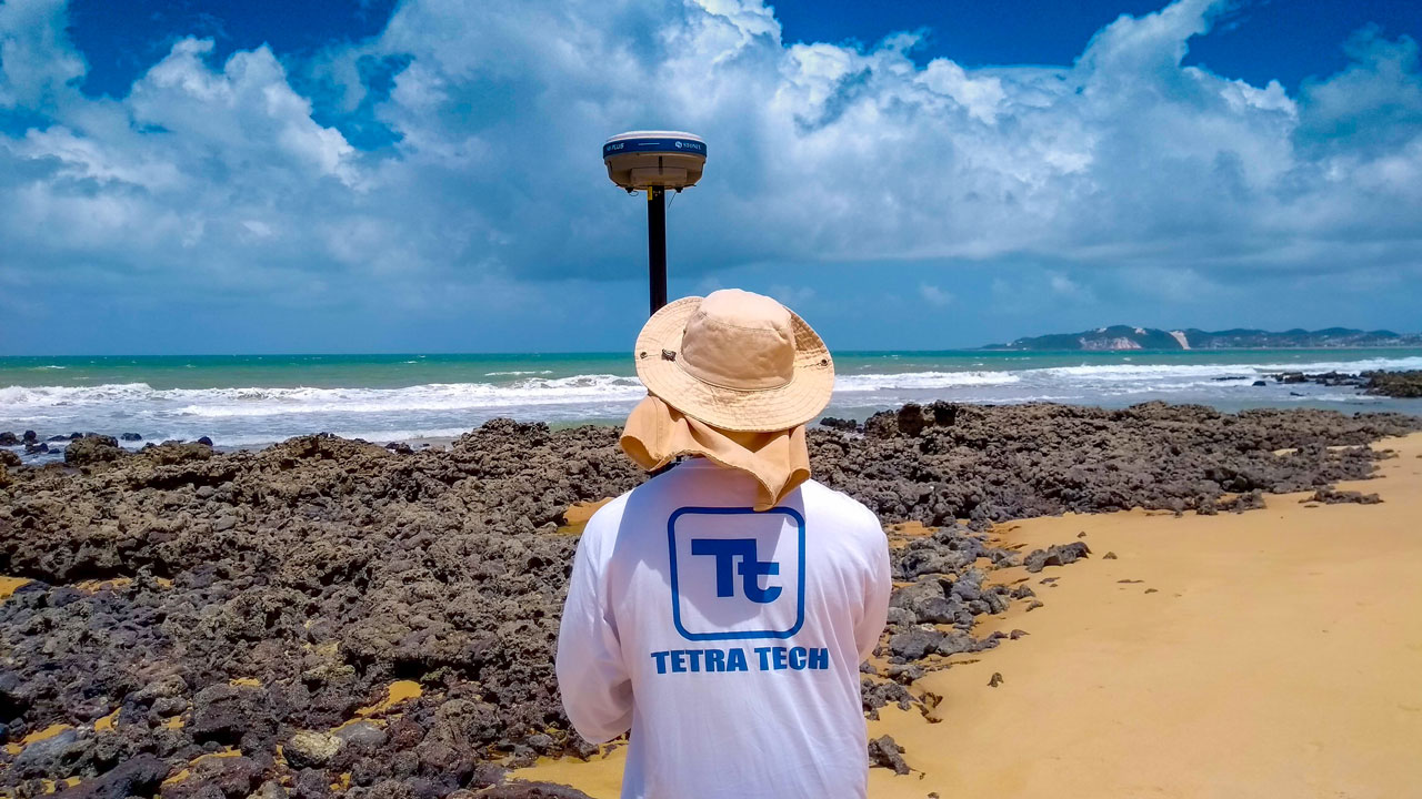 An employee in a Tetra Tech shirt and protective sun hat faces away from the camera, holding survey equipment and observing a stunning beach scene with a stretch of sand, rocks, ocean, and blue sky with fluffy white clouds