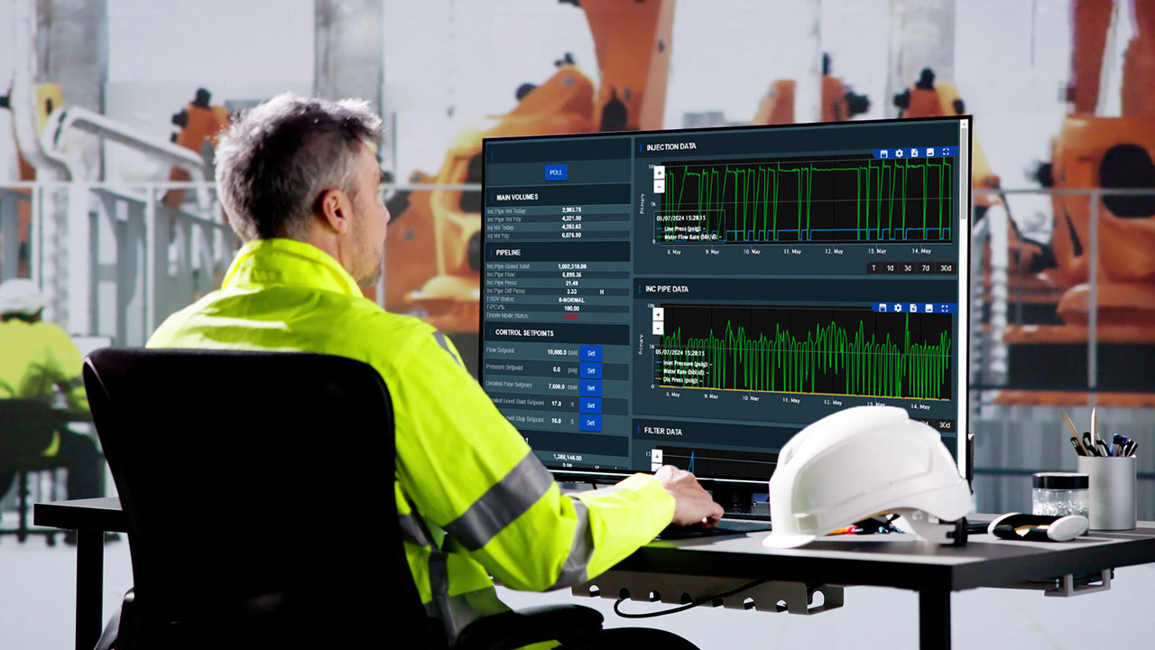 An engineer in a high-visibility jacket working on a computer with machinery in the background and a white hard hat on the desk