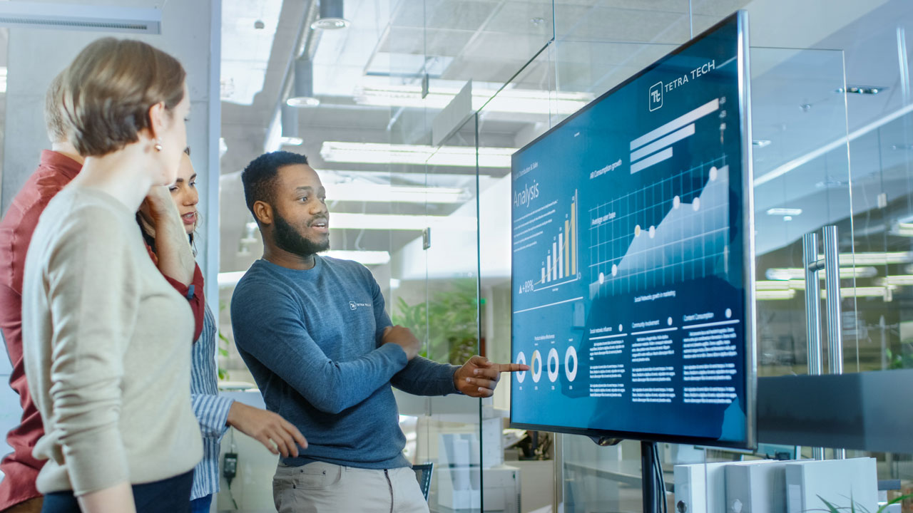 A group of professionals attentively watching a person presenting data and graphs on a large screen in a modern office