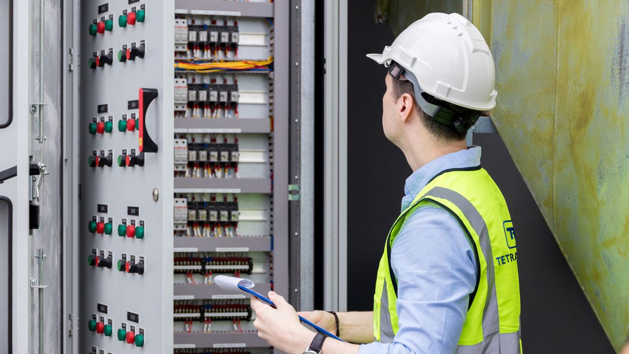 A technician wearing a white hard hat and high-visibility vest inspecting an electrical control panel and holding a clipboard