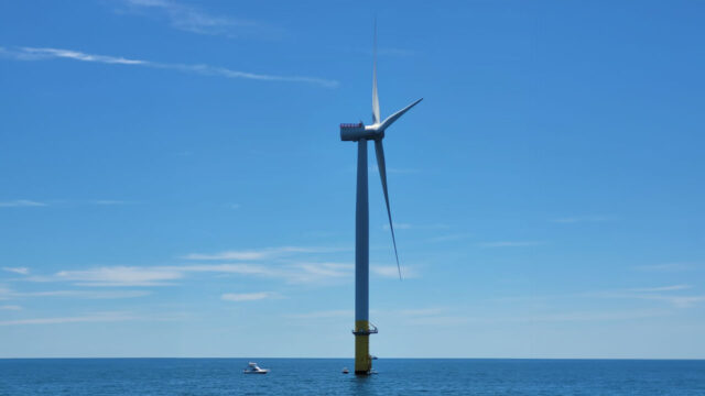 Close-up of an offshore wind turbine with boats in the background under a blue sky