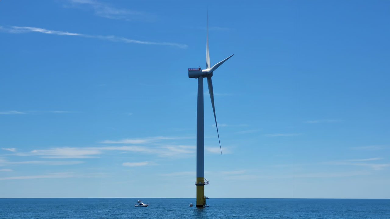 Close-up of an offshore wind turbine with boats in the background under a blue sky