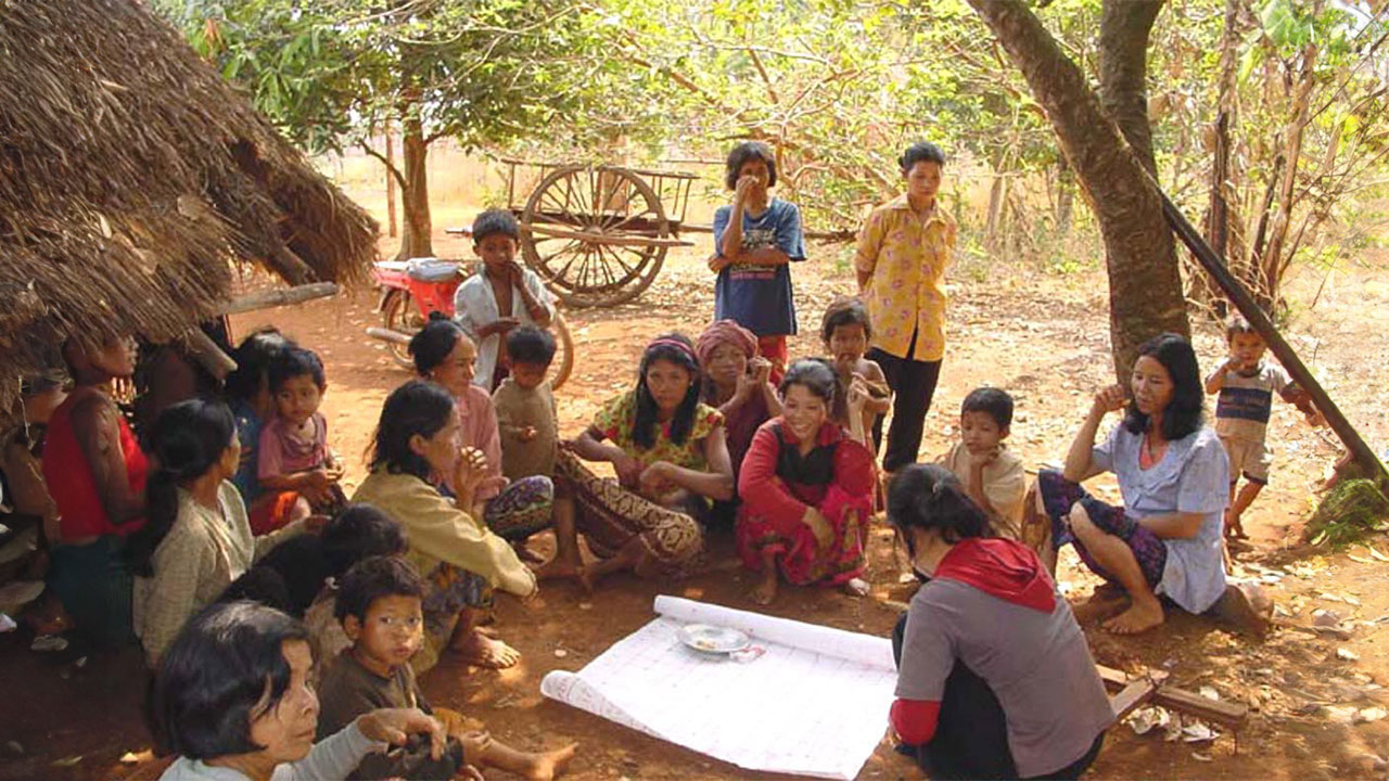 A community sits in a group on the floor around paper plans