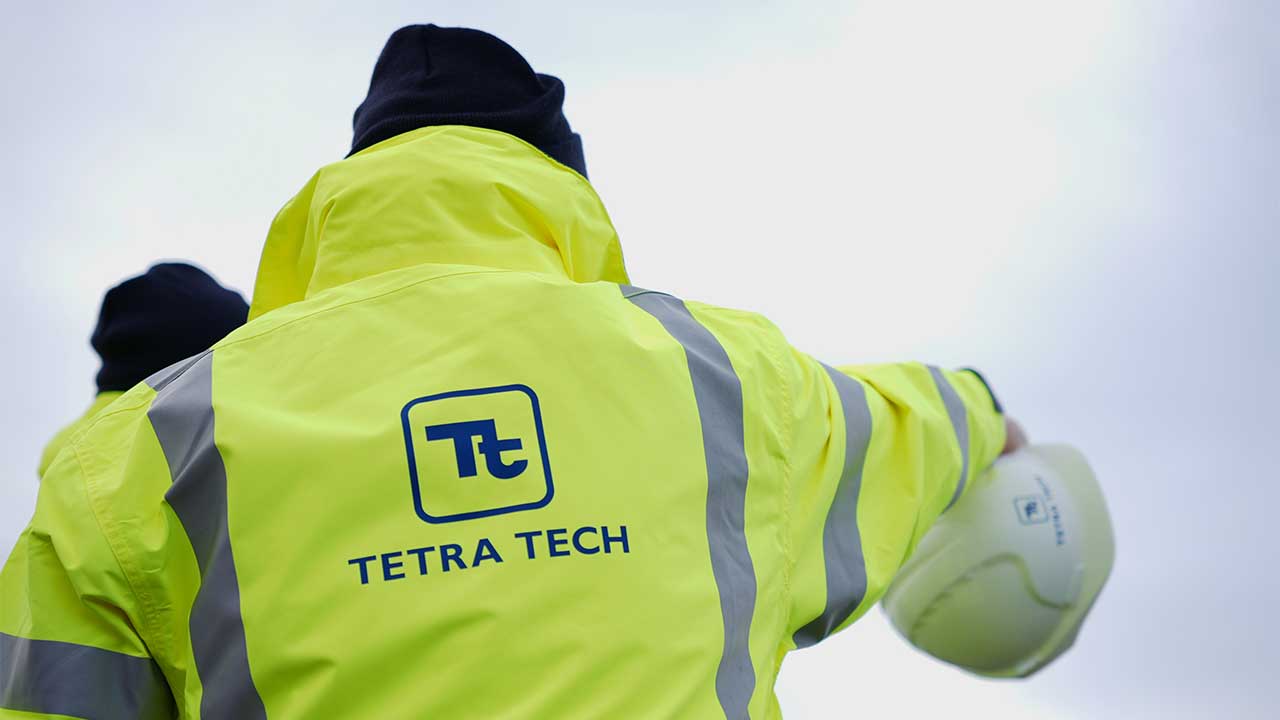 Person viewed from behind wearing a Tetra Tech branded high visibility jacket holding hard hat and pointing to horizon