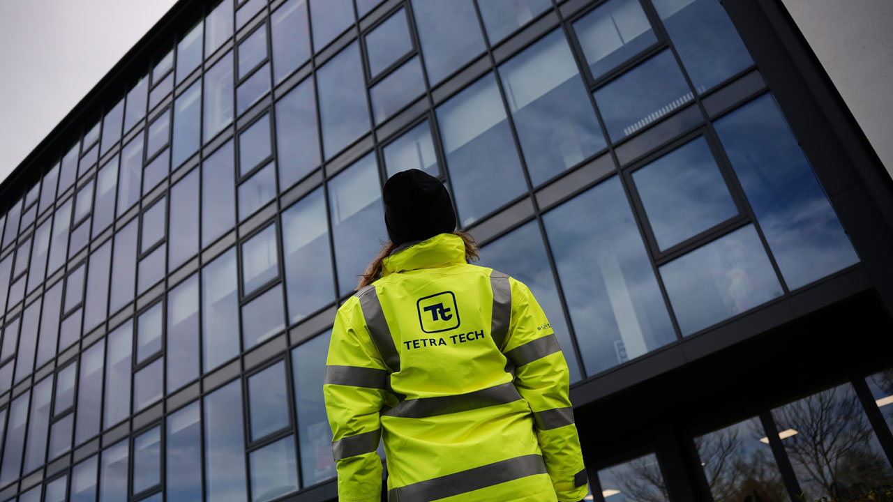 Person facing away from the camera wearing a high visibility outfit with a Tetra Tech logo on the back and a tall building in behind