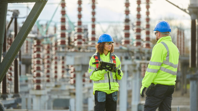 Two workers in high vis vest stand in front of electricity infrastructure