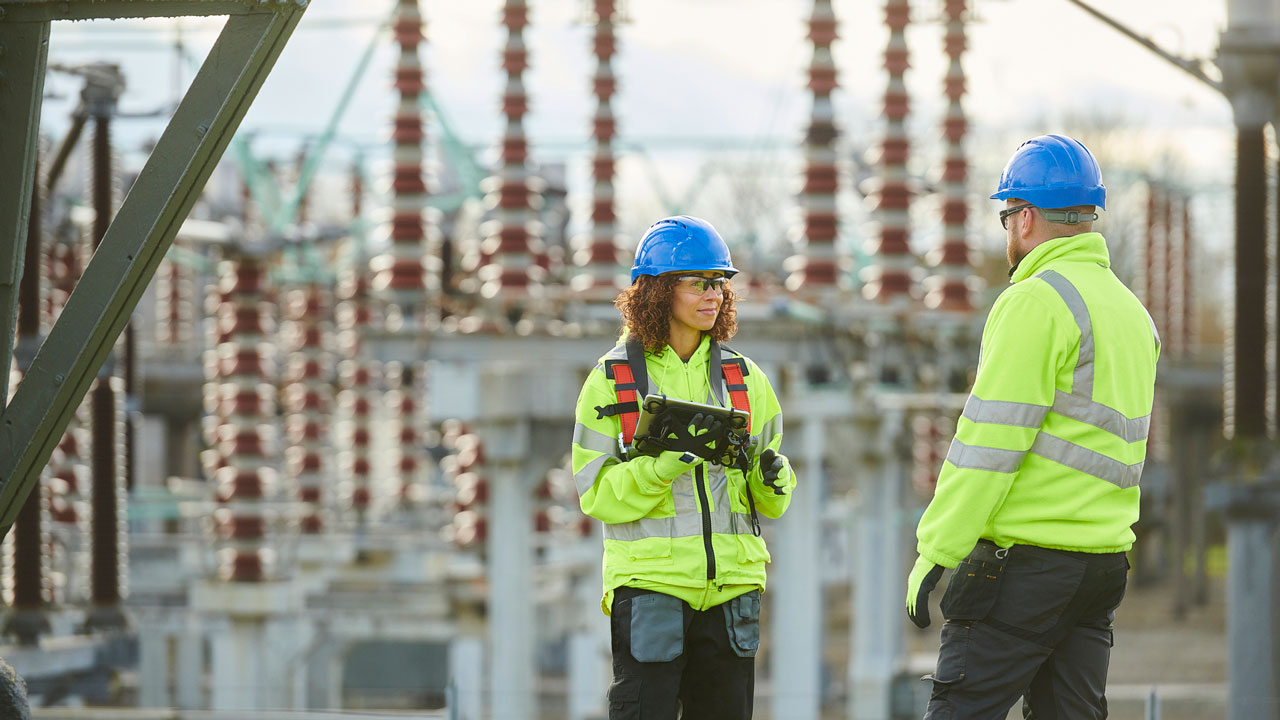 Two workers in high vis vest stand in front of electricity infrastructure