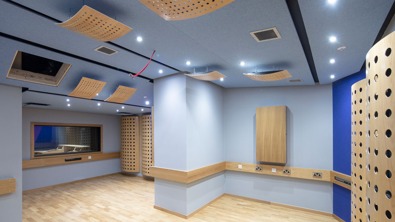 An interior view of a modern sound studio featuring acoustic panels on the ceiling, wooden accents, and a control room window