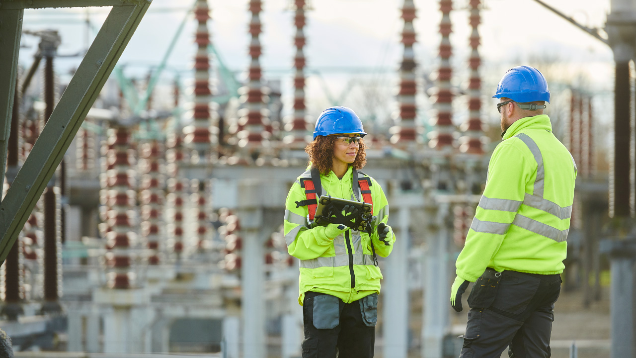 Two workers in safety gear discussing in an electrical substation, with power equipment in the background