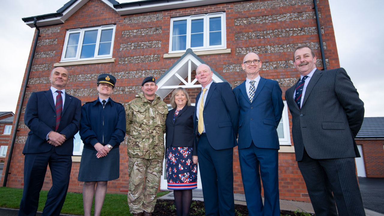 A group of seven individuals posing in front of a newly built brick house, smiling for the camera