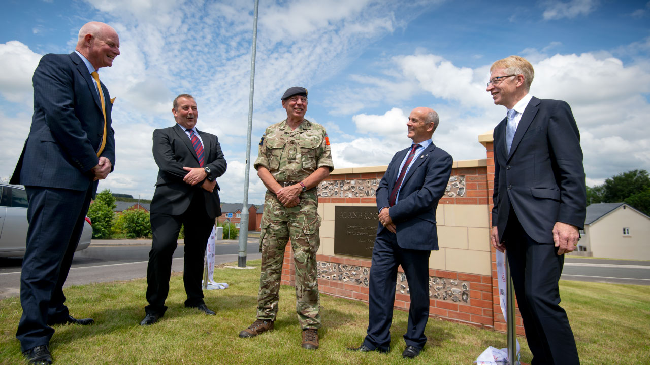 A group of five men, including a soldier in military uniform, gathered around a commemorative plaque, smiling and engaging in conversation. The background features a suburban setting with houses and a cloudy sky