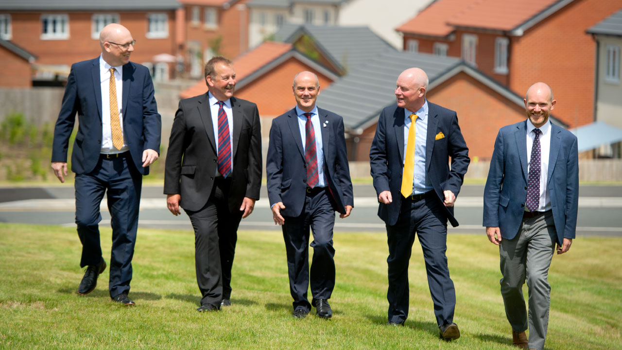 A group of five men in business attire walking together on a grassy area, with residential houses in the background