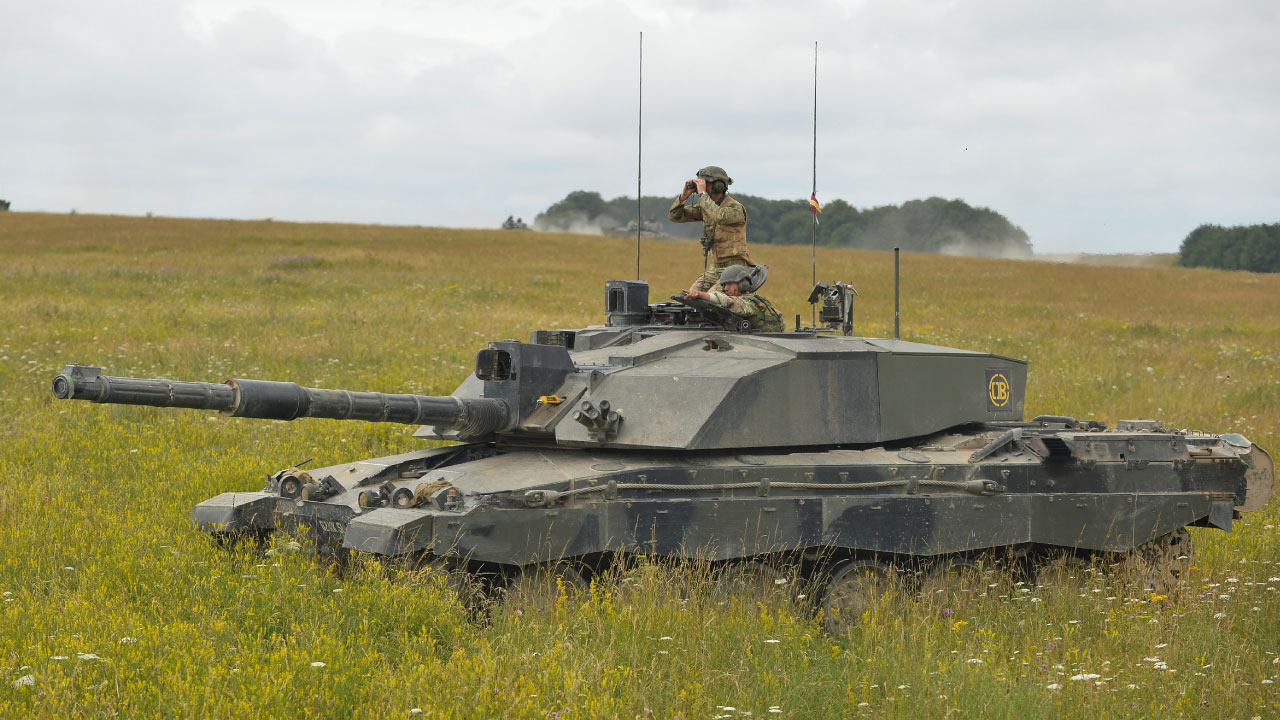 A military tank in a grassy field with soldiers on top, one using binoculars