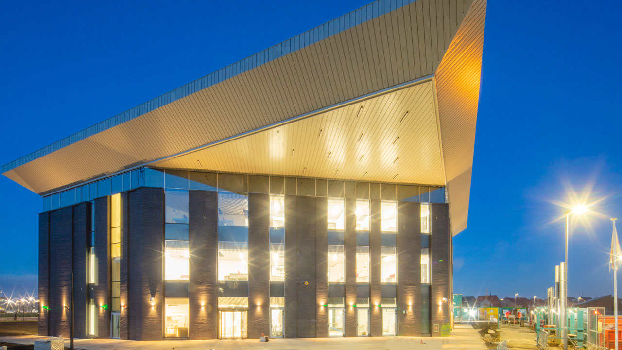 Multi-story building with angular roof, jutting awning, and many windows, illuminated against blue sky