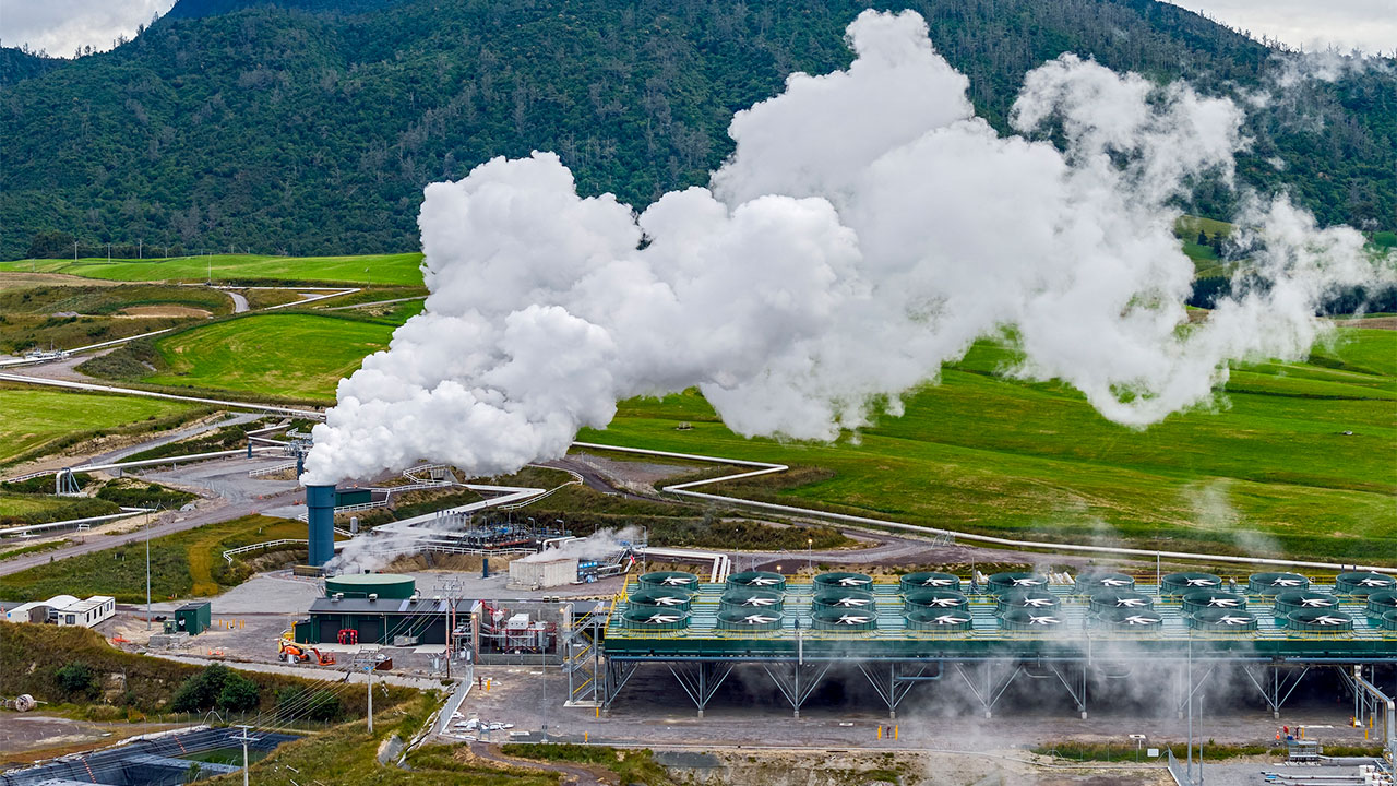 Aerial view of a geothermal power station with steam coming out of a stack and green fields and a mountain in the background