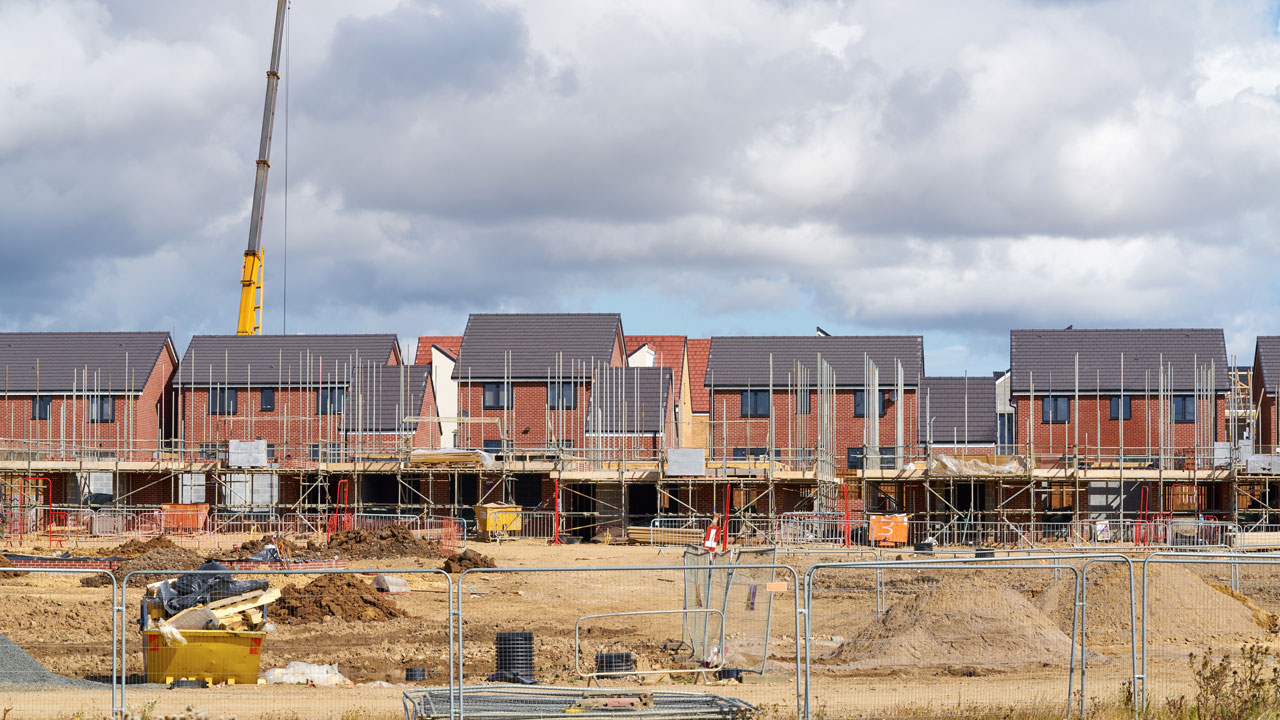 Construction fences and scaffolding in front of a row of red brick houses in the same style, with a large crane in the background