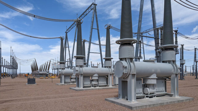 High-voltage substation equipment with large circuit breakers and insulators installed outdoors on a gravel yard, with transmission lines and steel support structures under a clear blue sky