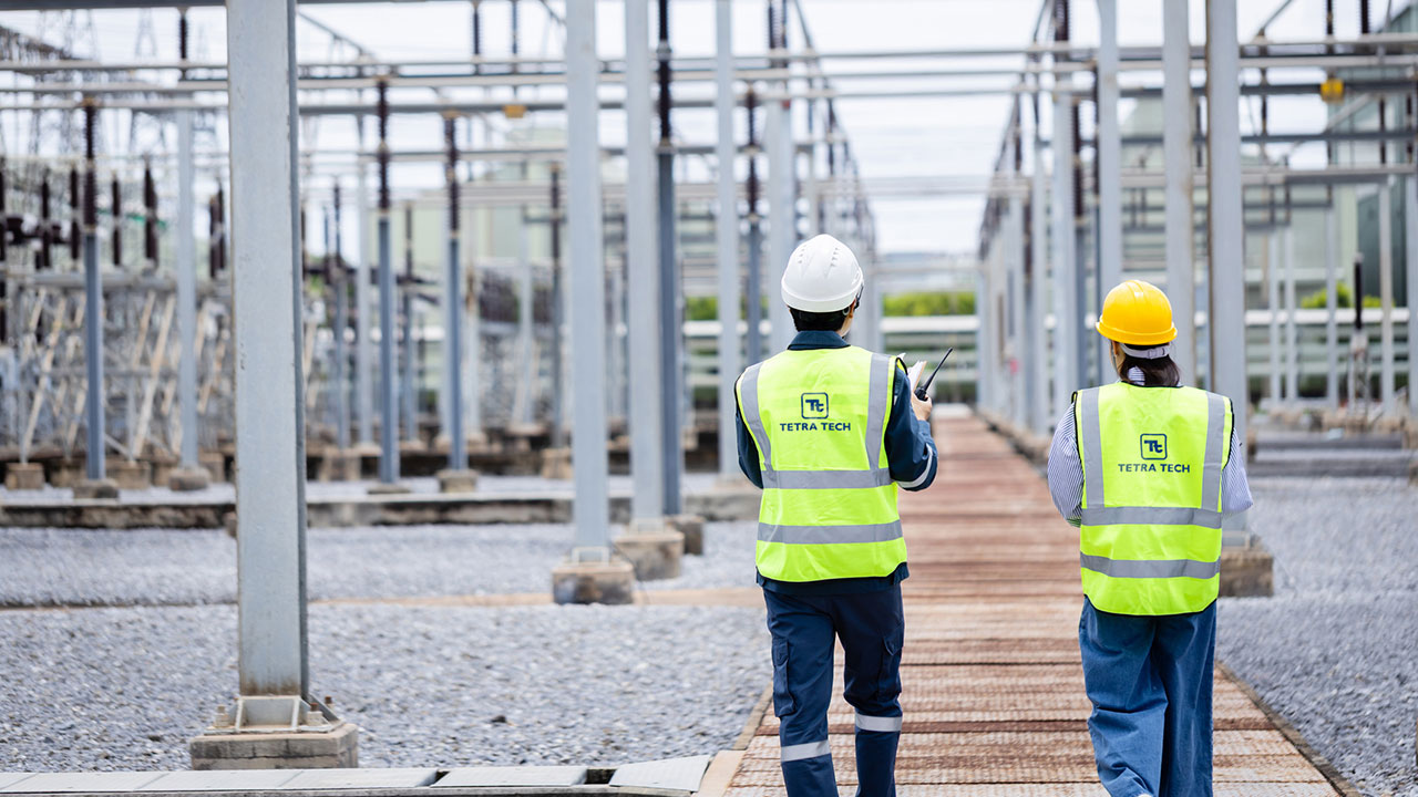 Two people in high visibility vests and hardhats walking through a substation