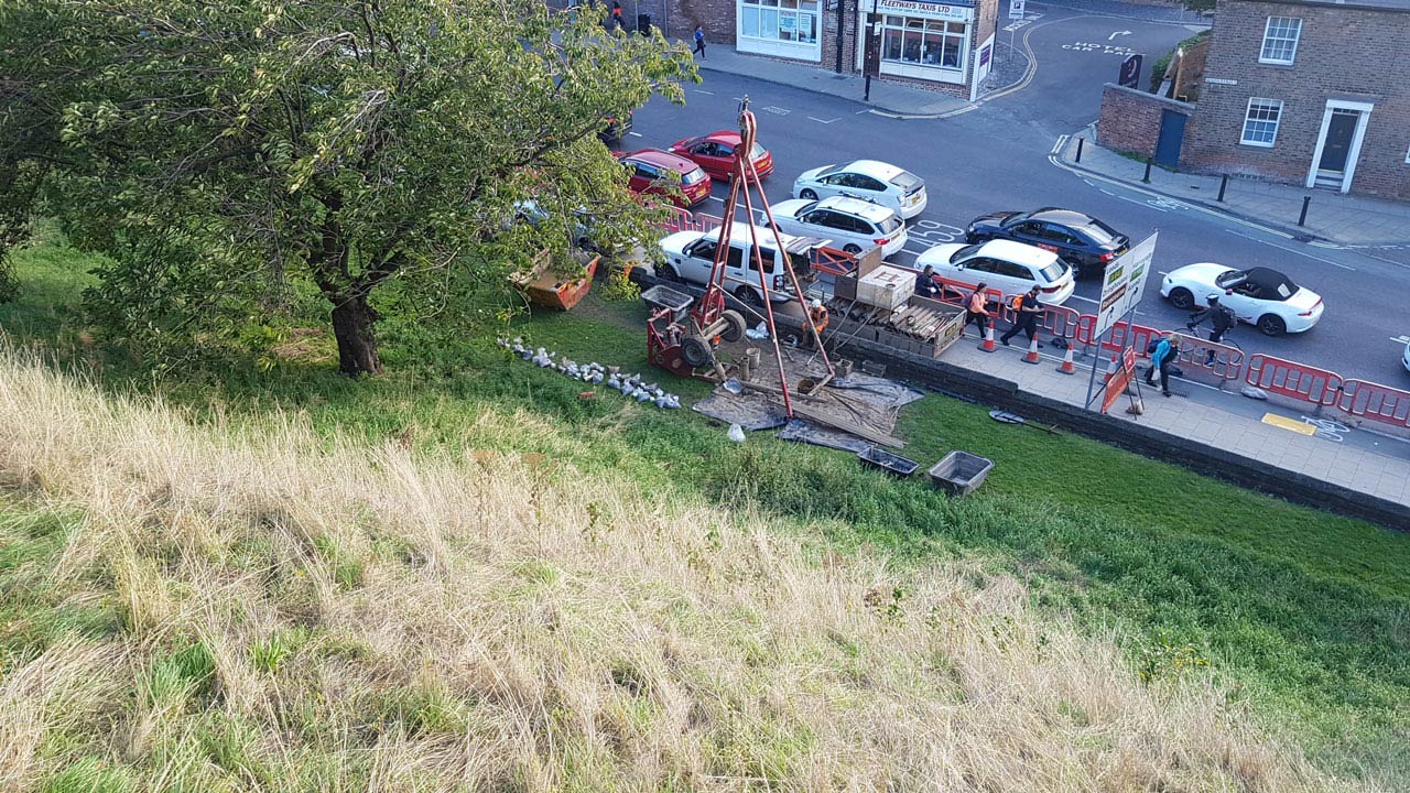 A construction site with machinery and workers near a grassy area, alongside a busy road with parked cars
