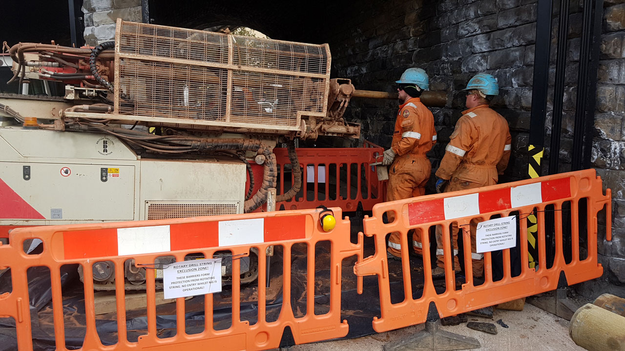 two construction workers in orange jumpsuits and hard hats operating machinery behind safety barriers in a stone archway