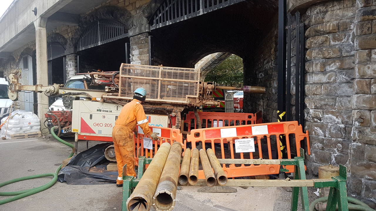 A construction worker in an orange jumpsuit operating machinery near a stone archway, surrounded by safety barriers and equipment