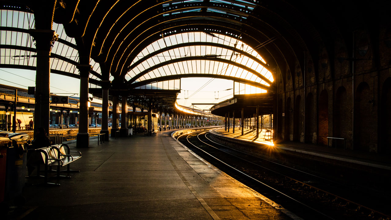 A train station platform at sunset, featuring arched glass roofing and illuminated tracks