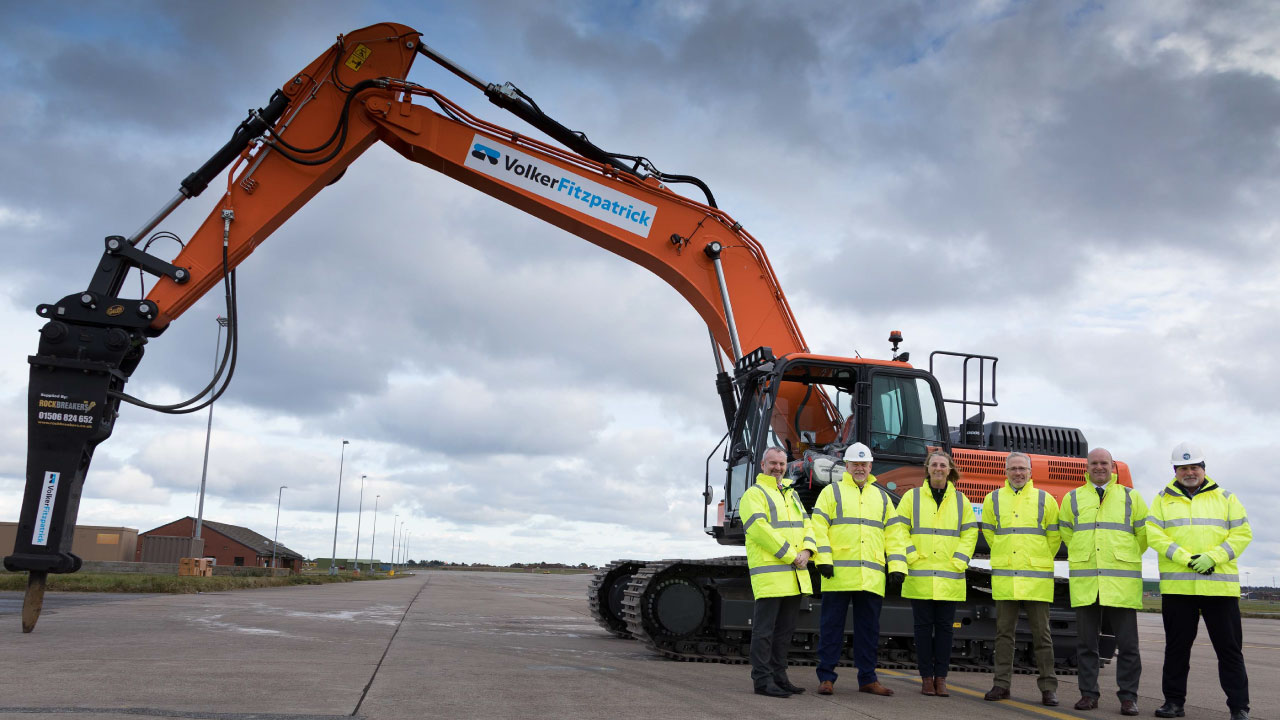A group of six individuals in high-visibility jackets and hard hats standing in front of a large excavator