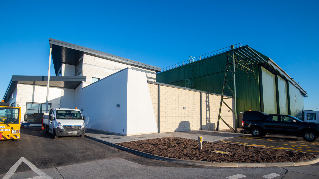 A modern industrial building with a green metal structure and a white facade, featuring parked vehicles in front
