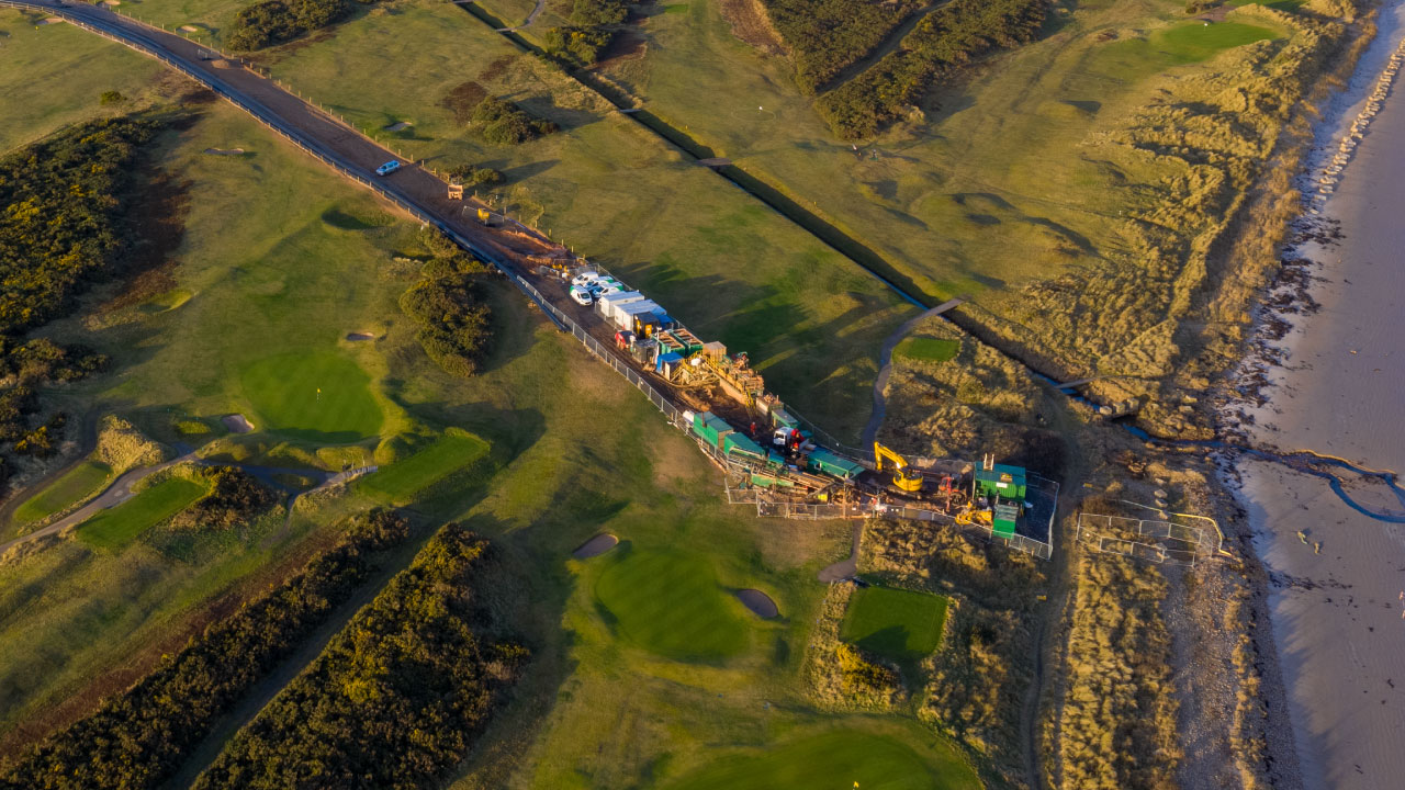 Aerial view of a construction site near a coastline, surrounded by green fields and golf courses