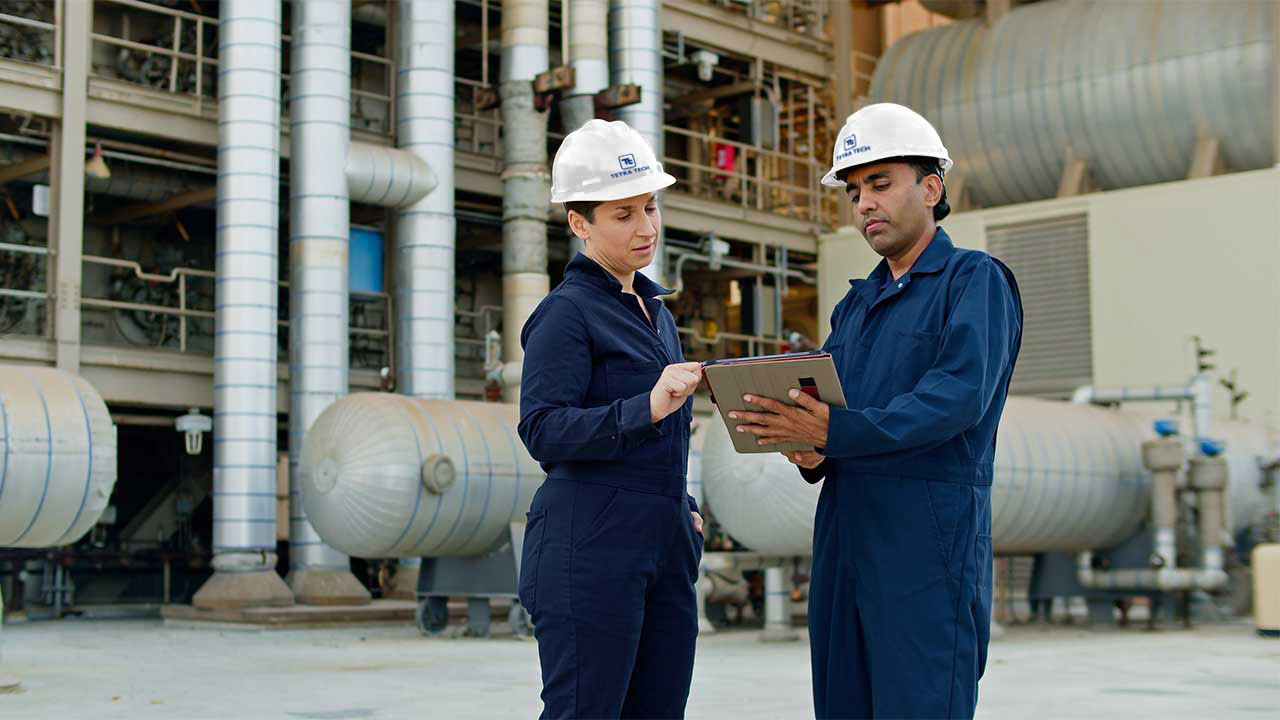 Still portrait of two industrial workers wearing hard hats and coveralls standing in front of a natural gas fired power station