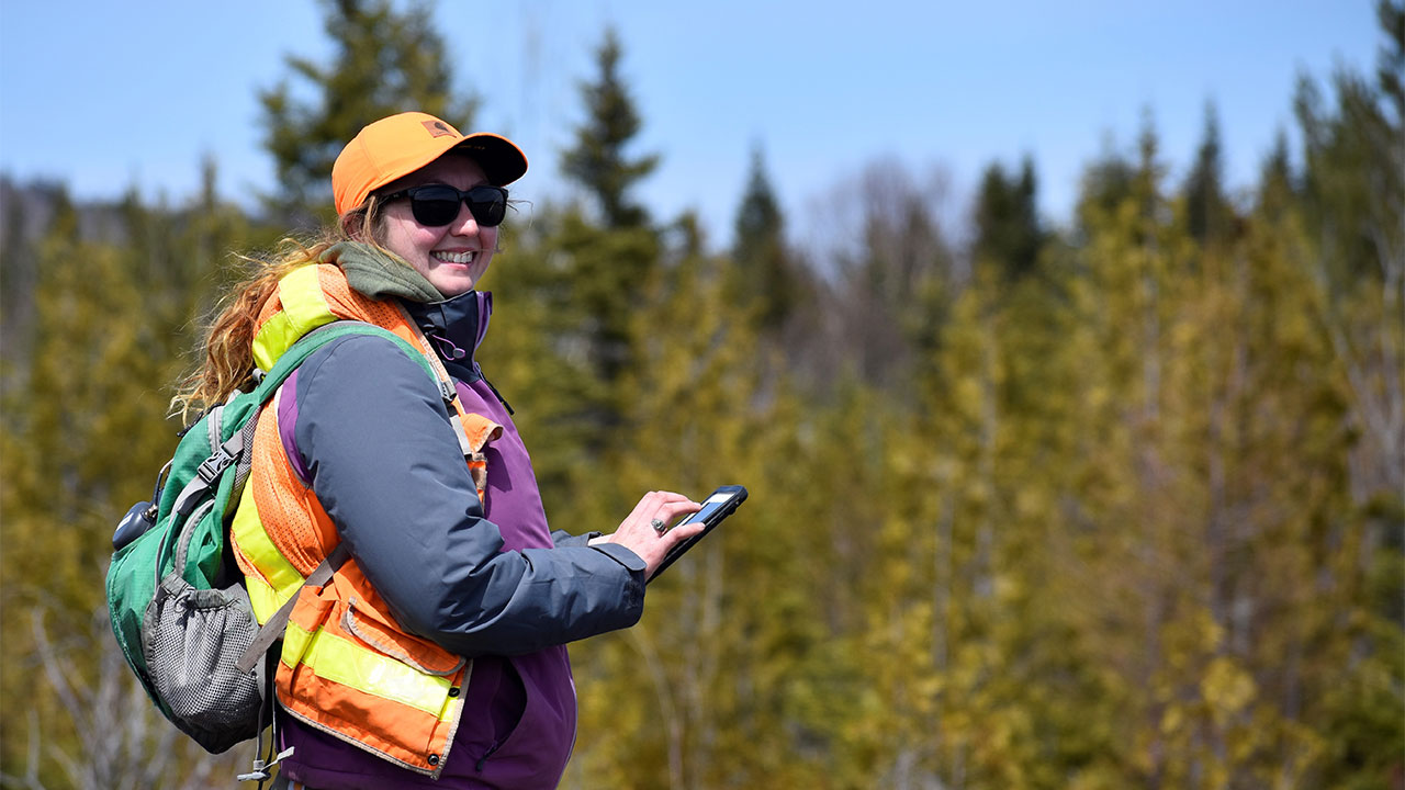 Field technician wearing high-visibility safety gear uses a handheld GPS device to collect data in a forested area