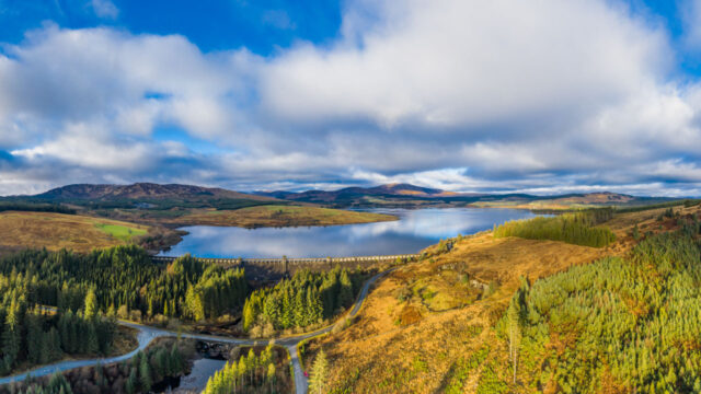 Aerial view of a scenic landscape featuring a lake, rolling hills, and a bridge crossing the water, surrounded by lush greenery and patches of brown land under a partly cloudy sky