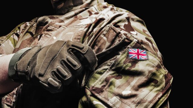 Close-up of a soldier's arm in camouflage uniform with a British flag patch, wearing a tactical glove, against a black background