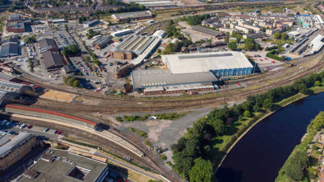 Aerial view of a large railway yard with multiple tracks, industrial buildings, parked cars, and a river curving along one side of the scene