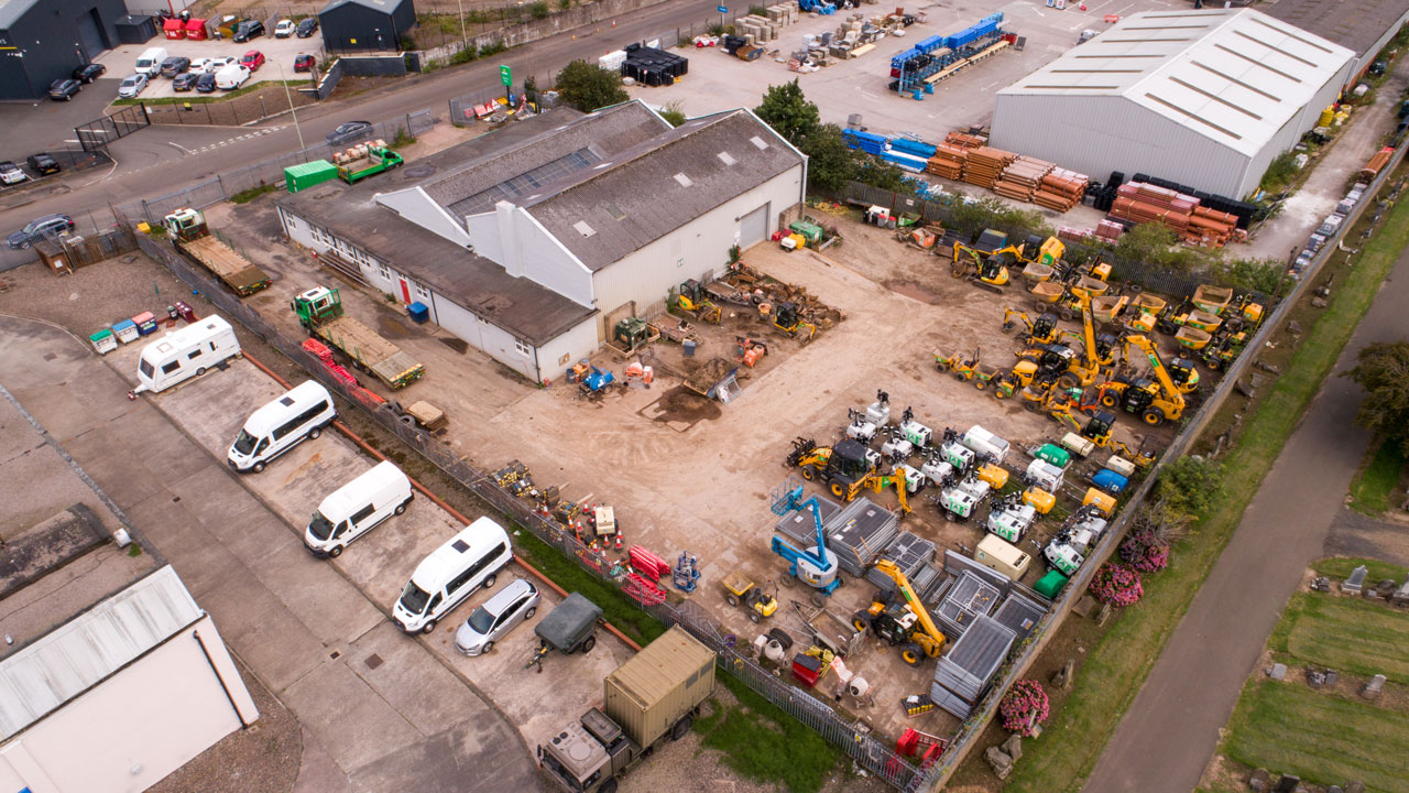 Aerial view of a construction yard filled with various construction vehicles and equipment, with a building in the center and a road nearby