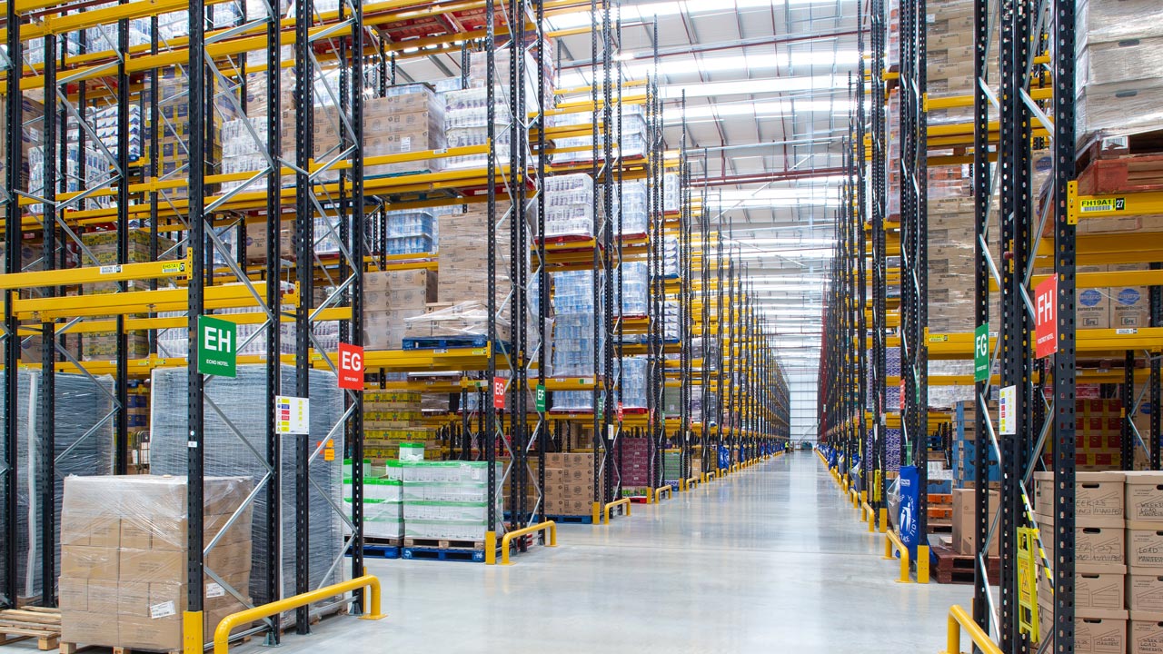 Interior of a high-bay warehouse with tall pallet racking filled with boxed goods and wide aisles
