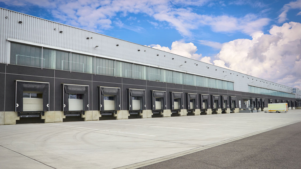 Large distribution warehouse with multiple loading bay doors and empty concrete yard under a blue sky