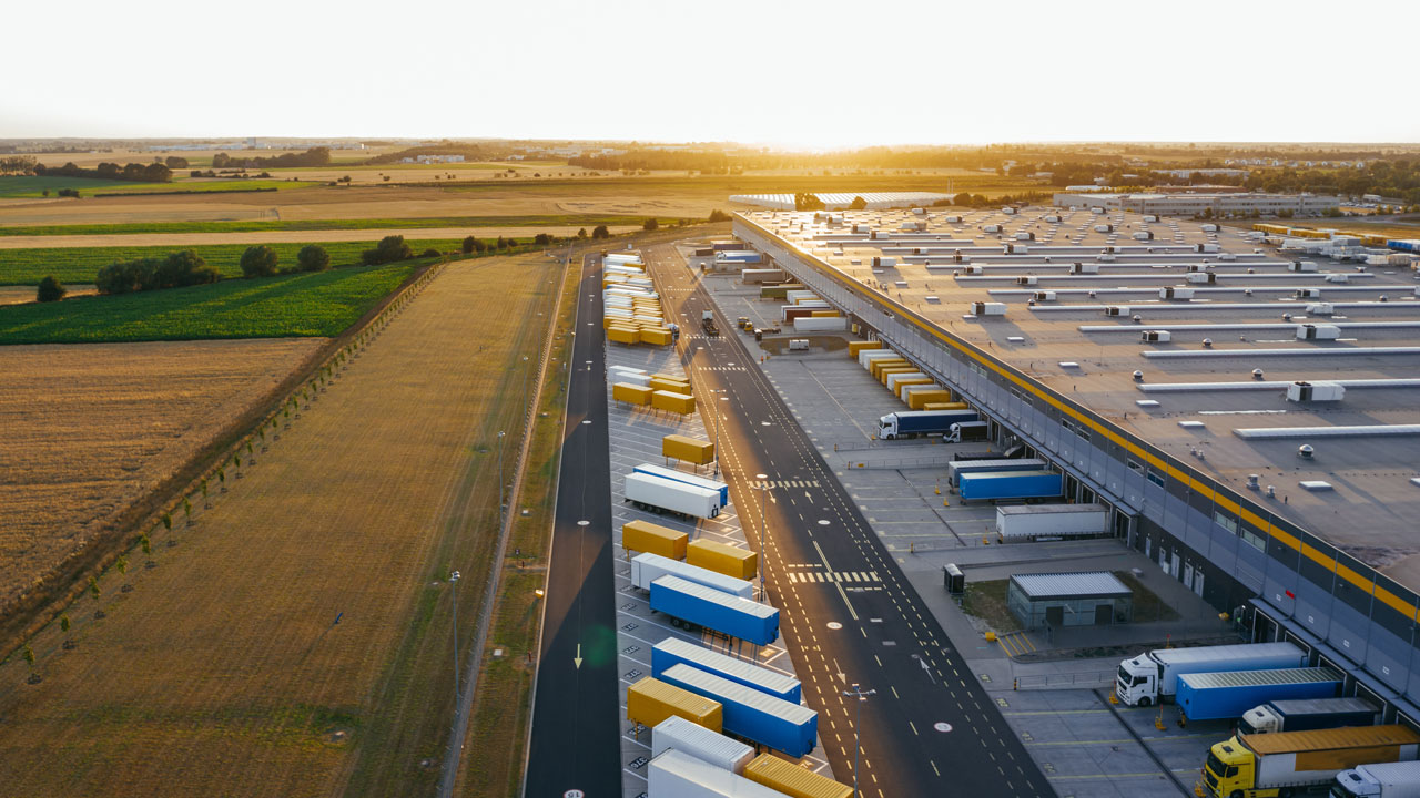 Aerial view of a large logistics distribution centre with multiple loading docks and parked delivery trailers at sunset